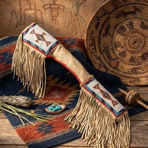 Peut inclure: Un fourreau en cuir orné de perles et de franges de style amérindien, avec un accent rouge, repose sur un tapis bleu et orange à motifs. Un panier tissé et un petit plat de pierres turquoises sont également visibles, ainsi qu'un paquet de sauge.