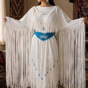 May include: A white long-sleeved dress featuring extensive fringe detailing along the sleeves and hem. The garment includes a bright blue waist belt adorned with white quill-like accents and small blue beads, with matching beadwork on the bodice.