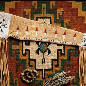 Puede incluir: Una vaina de cuero con cuentas nativa americana con flecos. La vaina es de color canela con secciones de cuentas blancas con diseños geométricos en verde, rojo y negro. El fondo es una alfombra estampada con colores tierra.
