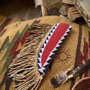 May include: A handcrafted, beaded knife sheath with a red center and white and blue triangle border. The sheath has a fringed leather backing. A pair of feathers and turquoise beads are also visible.