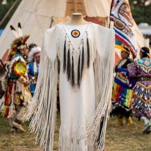 May include: A white fringed dress with a round neckline and a colorful circular emblem. The dress has long fringe sleeves and a fringed hemline. The dress is displayed on a mannequin, with people in traditional Native American attire in the background.