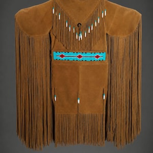 Puede incluir: Prenda de ante marrón con largos flecos. La prenda presenta una banda decorativa con detalles turquesa y rojos. El escote y los hombros también están adornados con flecos y pequeñas cuentas. El diseño sugiere una influencia nativa americana.