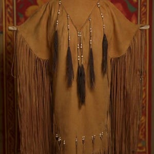 May include: A tan suede poncho with long fringe and decorative beaded accents. The poncho features a V-neckline and is adorned with beaded strands and dark brown tassels. The fringe cascades down the sides, creating a flowing silhouette.