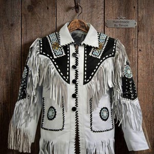 May include: A white jacket featuring black panels on the chest and sleeves, adorned with long white fringe, black whipstitch detailing, and circular beaded accents. The front includes five black buttons and a collar, with intricate geometric beadwork.