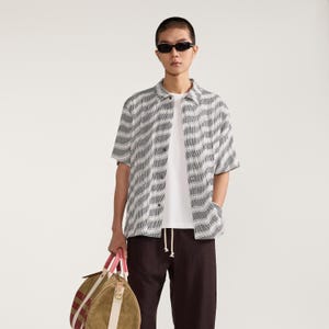 May include: A short-sleeved button-down shirt with a grey and white wavy pattern, worn over a plain white t-shirt. The outfit includes dark brown trousers with a drawstring waist and a tan duffel bag featuring pink handles and red striped accents.