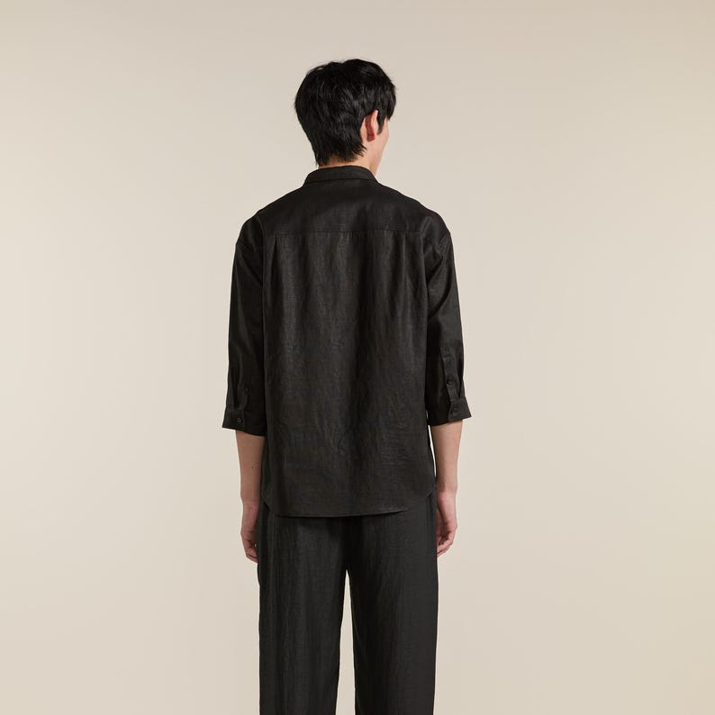Puede incluir: Una camisa negra de botones con mangas tres cuartos y cuello, combinada con pantalones negros a juego. La camisa presenta un canes&uacute; en la espalda y un dobladillo recto, mostrando un acabado de tela texturizada en toda la prenda.