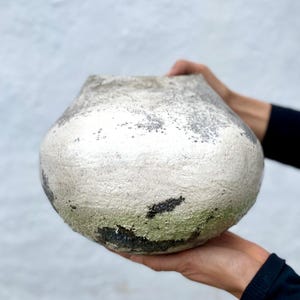 Pode incluir: Um vaso de cerâmica arredondado com um corpo largo e texturizado, apresentando um acabamento manchado em tons de branco sujo, cinza e carvão. A superfície tem uma aparência áspera e desgastada com manchas escuras e um tom verde na base.