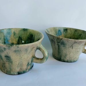 Puede incluir: Dos tazas de cerámica con un borde ancho y acampanado y una pequeña asa lateral. La superficie presenta un esmalte moteado con tonos mezclados de crema, verde salvia y toques de azul, creando una apariencia texturizada y variada en el exterior.