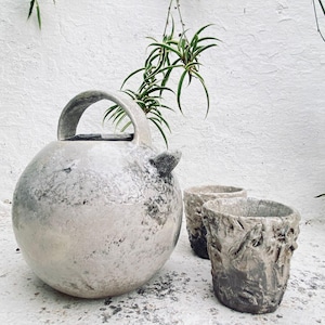 Puede incluir: Un recipiente de cerámica esférico con asa curva y un pequeño pico lateral, acompañado de dos tazas de cerámica cilíndricas y texturizadas. Todos los objetos presentan un acabado moteado y desgastado en gris y blanco roto.