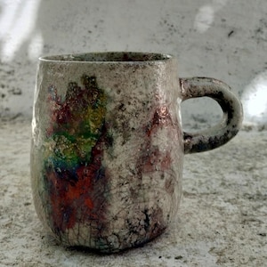 Puede incluir: Una taza de cerámica con una superficie texturizada y moteada en color blanco roto, con un acabado de esmalte craquelado. El exterior muestra una mancha abstracta y vibrante de colores iridiscentes, incluyendo verde, azul y rojo, con asa.