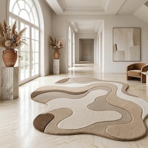 May include: An area rug with an irregular, organic shape featuring layered, flowing sections in shades of cream, beige, tan, and dark brown. The rug has a textured surface with defined, curved borders separating each color block across the floor.