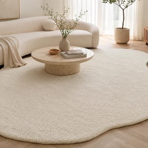 May include: An off-white, plush area rug with an irregular, organic, amoeba-like shape. The rug features a thick, textured pile surface and is positioned on a light-colored wooden floor, extending underneath a round, light-toned stone coffee table.