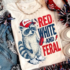 Könnte beinhalten: Cremefarbenes T-Shirt mit einem Waschbären, der eine rot-weiß Baseballkappe mit dem Wort "Merica" trägt. Das Shirt hat die Worte "Red White and Feral" in Rot und Blau. Der Waschbär hält einen Hotdog und trägt weiße Clogs.