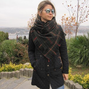 May include: A woman wearing a black quilted jacket with a plaid scarf and mirrored sunglasses. She is standing on a stone path with a view of a city in the background.