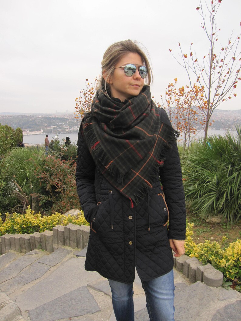 May include: A woman wearing a black quilted jacket with a plaid scarf and mirrored sunglasses. She is standing on a stone path with a view of a city in the background.