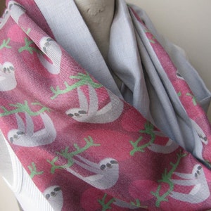 May include: A gray and pink infinity scarf with a repeating pattern of sloths hanging from branches. The sloths are white with black eyes and noses.