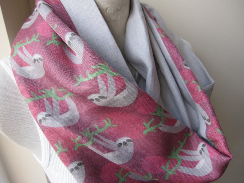 May include: A gray and pink infinity scarf with a repeating pattern of sloths hanging from branches. The sloths are white with black eyes and noses.
