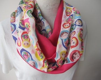 May include: A white infinity scarf with a pink lining and a colorful pattern of Russian nesting dolls. The dolls are depicted with smiling faces and floral designs.