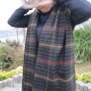 May include: A black and brown plaid scarf with red and tan stripes. The scarf is worn by a person with long, blonde hair.