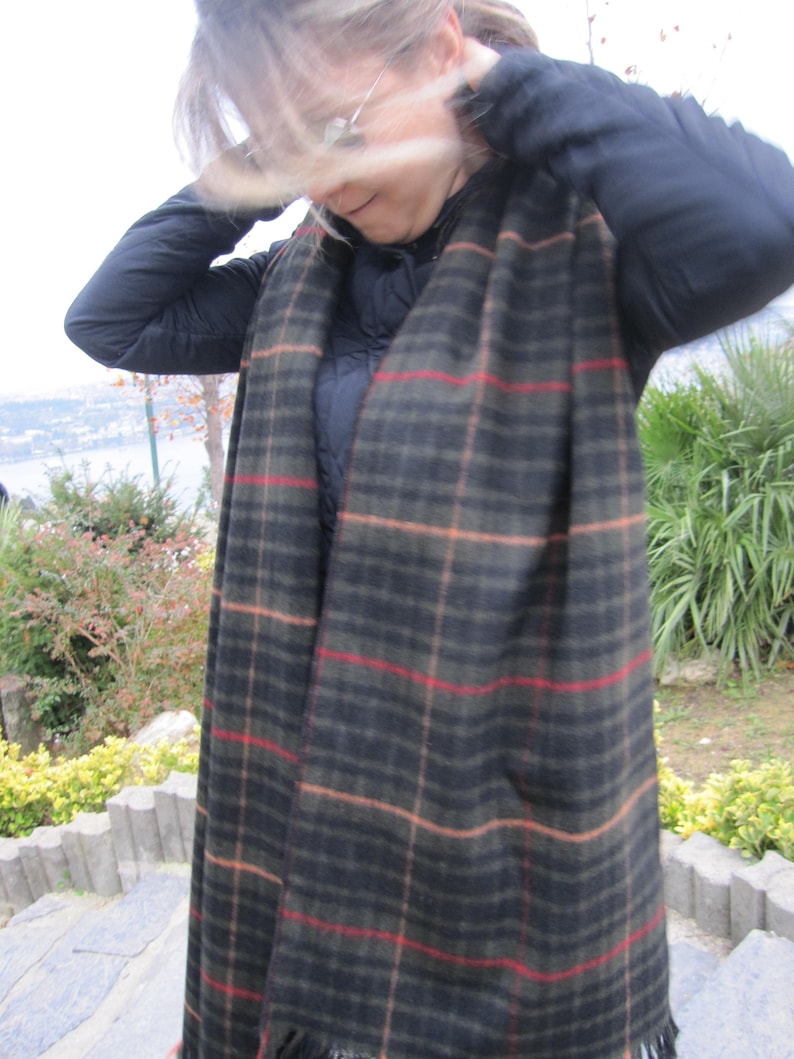 May include: A black and brown plaid scarf with red and tan stripes. The scarf is worn by a person with long, blonde hair.