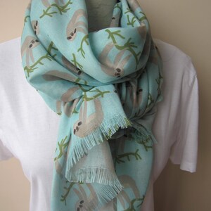 May include: A light blue scarf with a fringe border features a repeating pattern of sloths hanging from branches. The sloths are a light brown color and the branches are green.