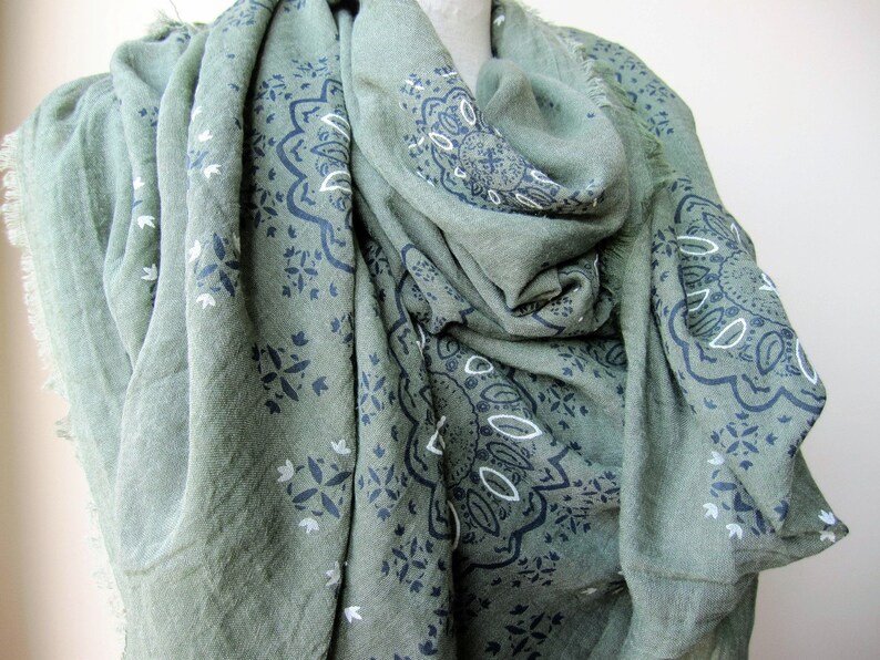 May include: A green scarf with a black and white paisley pattern. The scarf is made of a soft, lightweight fabric and has a fringed edge.