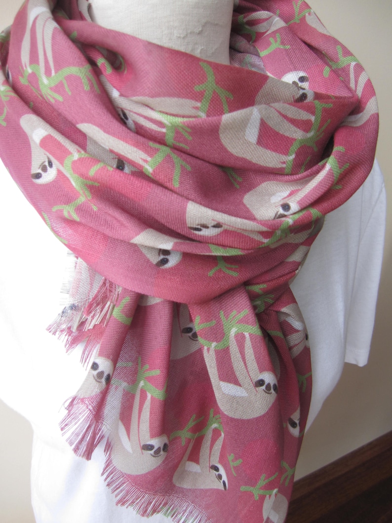 May include: A pink scarf with a pattern of sloths hanging from branches. The scarf has a fringed edge.