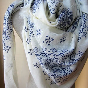 May include: White scarf with a blue paisley pattern. The scarf is made of a soft, lightweight fabric and has a fringed edge.