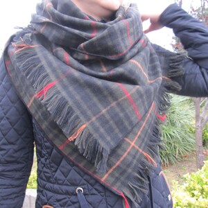 May include: A black, green, orange, and red plaid scarf with fringe. The scarf is worn around the neck of a person wearing a black quilted jacket.