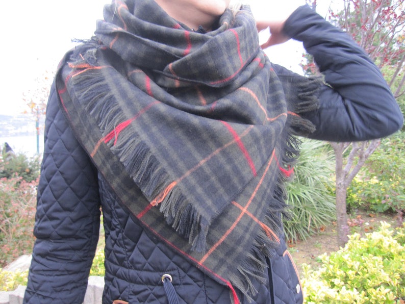 May include: A black, green, orange, and red plaid scarf with fringe. The scarf is worn around the neck of a person wearing a black quilted jacket.