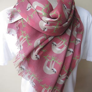 May include: A pink scarf with a sloth print. The sloths are beige and are hanging from green branches. The scarf has a fringed edge.