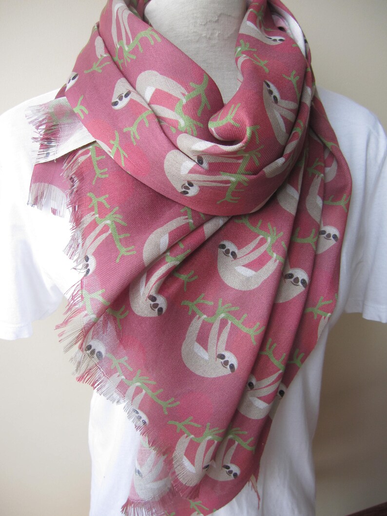 May include: A pink scarf with a sloth print. The sloths are beige and are hanging from green branches. The scarf has a fringed edge.