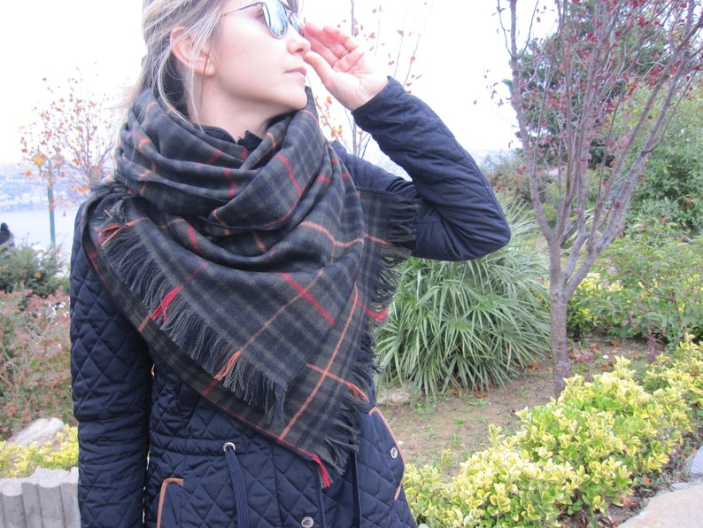 May include: A woman wearing a black quilted jacket with a large black and brown plaid scarf with fringe. The scarf is wrapped around her neck and shoulders.