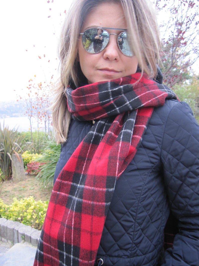 May include: A woman wearing a navy blue quilted jacket with a red and black plaid scarf. She is wearing sunglasses and has long blonde hair.