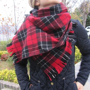 May include: A red and black plaid scarf with fringe edges. The scarf is worn around the neck of a person wearing a black quilted jacket.