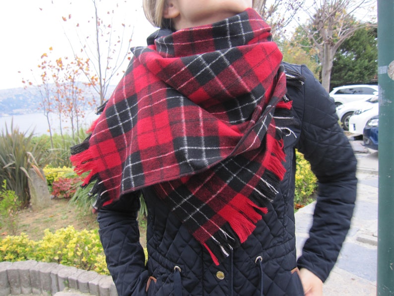May include: A red and black plaid scarf with fringe edges. The scarf is worn around the neck of a person wearing a black quilted jacket.