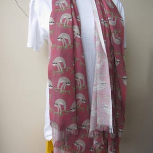 May include: A pink and white scarf with a sloth pattern. The scarf has a fringed edge and is made of a soft, lightweight fabric.