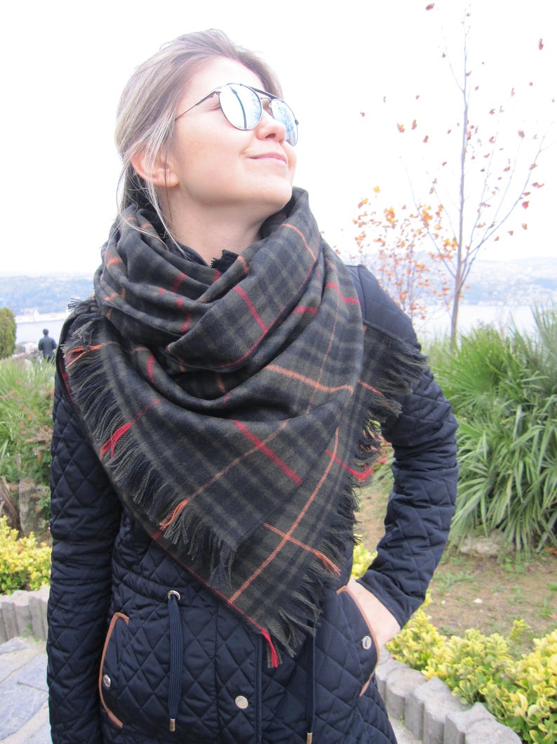 May include: A woman wearing a black quilted jacket with a brown trim and a black and green plaid scarf with fringe. She is wearing mirrored sunglasses and looking up.