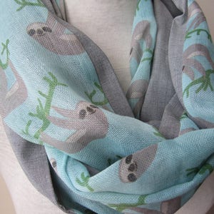May include: A light blue and gray infinity scarf with a sloth print. The scarf features sloths hanging from branches on a light blue background. The reverse side of the scarf is gray.