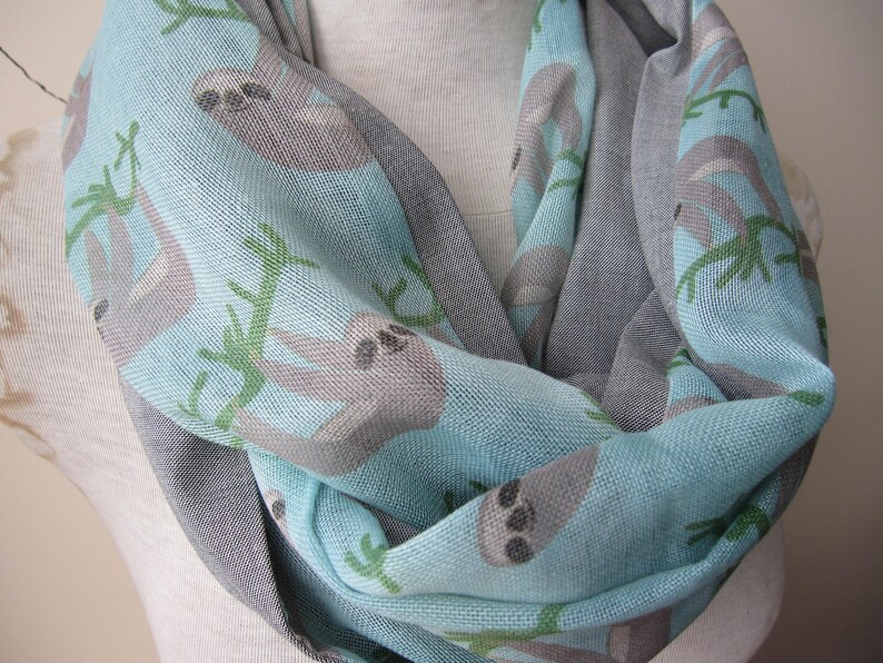 May include: A light blue and gray infinity scarf with a sloth print. The scarf features sloths hanging from branches on a light blue background. The reverse side of the scarf is gray.