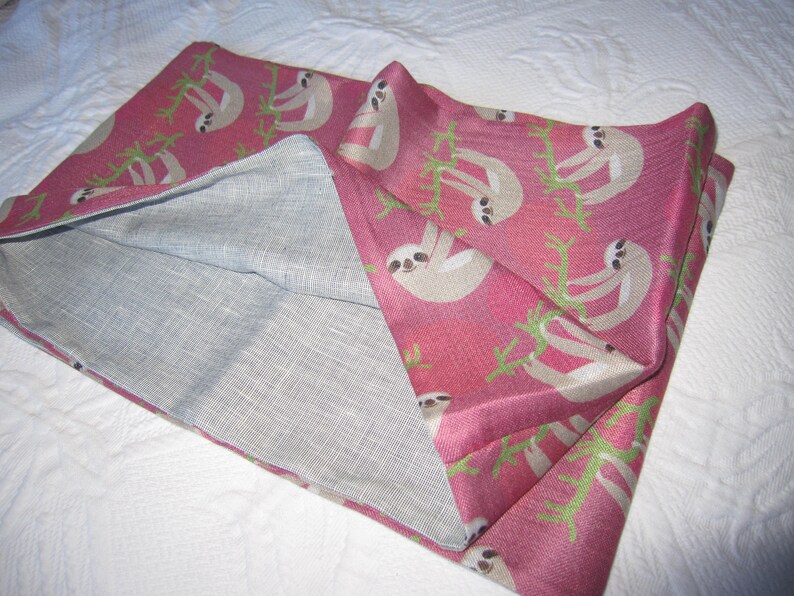 May include: A pink fabric pillowcase with a white sloth pattern. The pillowcase is folded in half, showing the inside of the fabric, which is a light gray.