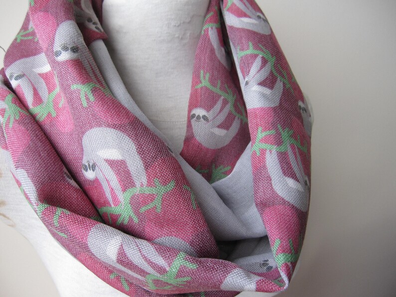May include: A pink and gray infinity scarf with a repeating pattern of sloths on branches. The sloths are white and have black eyes.