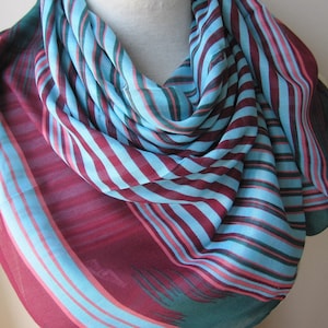May include: A striped scarf in shades of teal, burgundy, and pink. The scarf is made of a lightweight fabric and has a flowing drape.