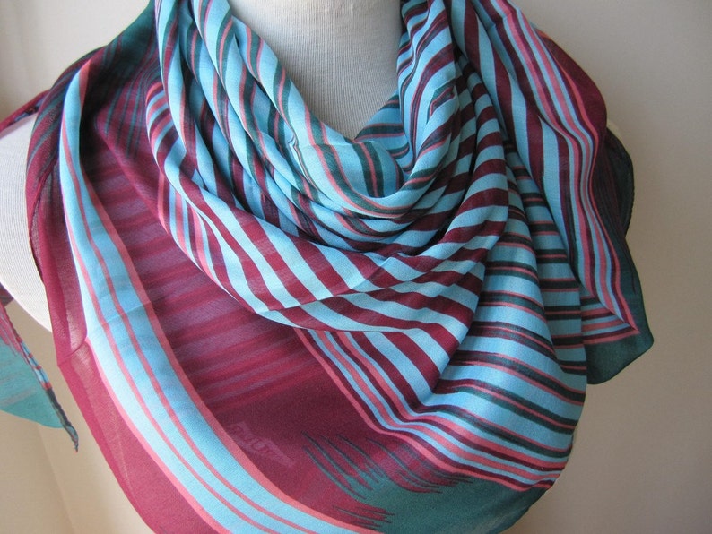 May include: A striped scarf in shades of teal, burgundy, and pink. The scarf is made of a lightweight fabric and has a flowing drape.