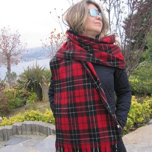 May include: A woman wearing a black jacket and a red and black plaid scarf with fringe. The scarf is wrapped around her neck and tied in a knot at the front. She is wearing sunglasses and looking to the right.