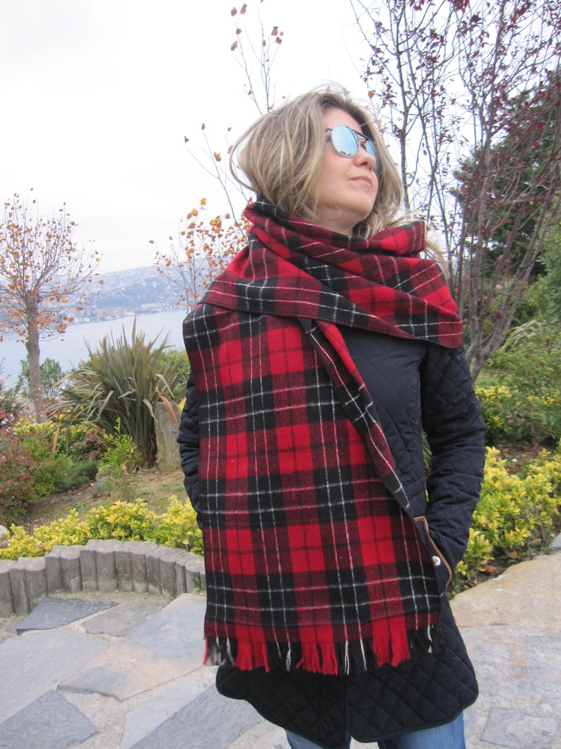 May include: A woman wearing a black jacket and a red and black plaid scarf with fringe. The scarf is wrapped around her neck and tied in a knot at the front. She is wearing sunglasses and looking to the right.