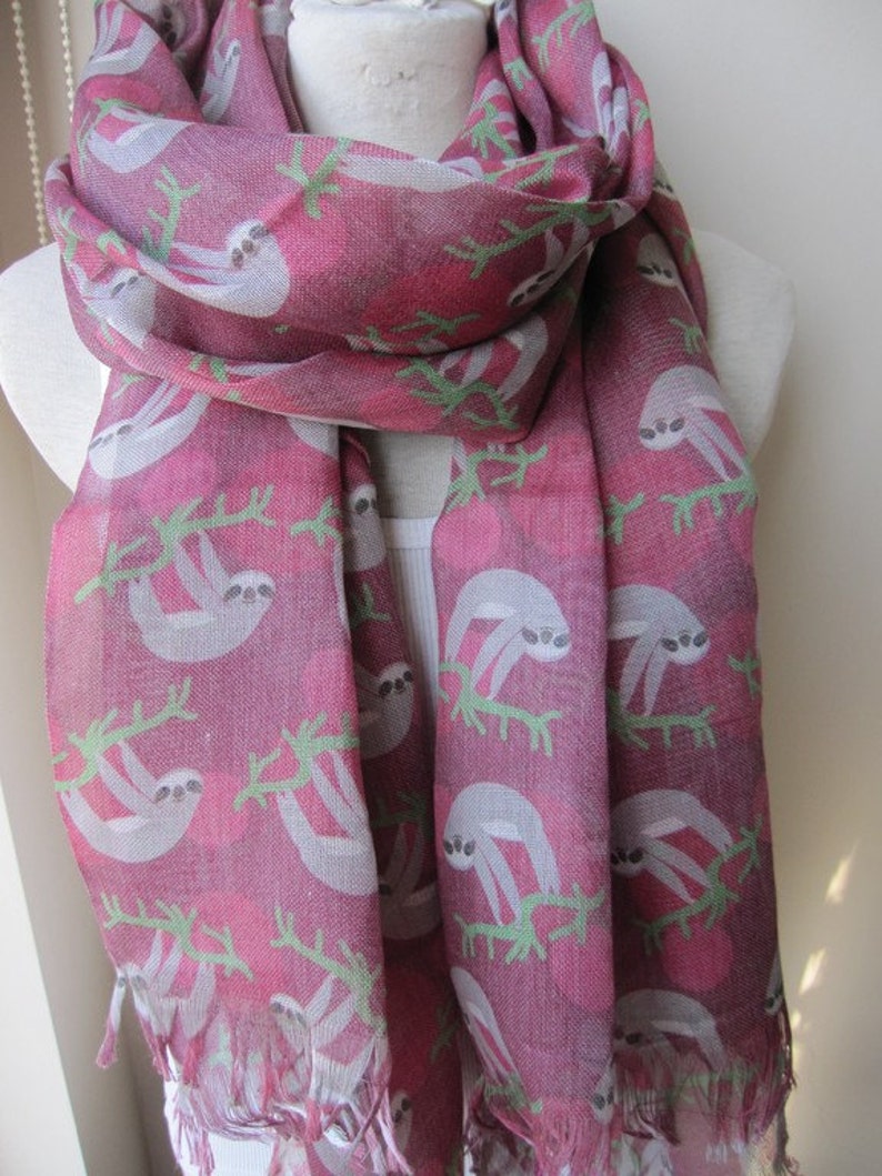 May include: A pink and gray scarf with a sloth pattern. The scarf has a fringe border and is made of a soft, lightweight fabric.