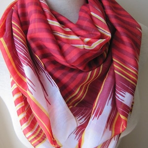 May include: A red and yellow striped scarf with a white and red geometric pattern. The scarf is folded and draped over a mannequin.