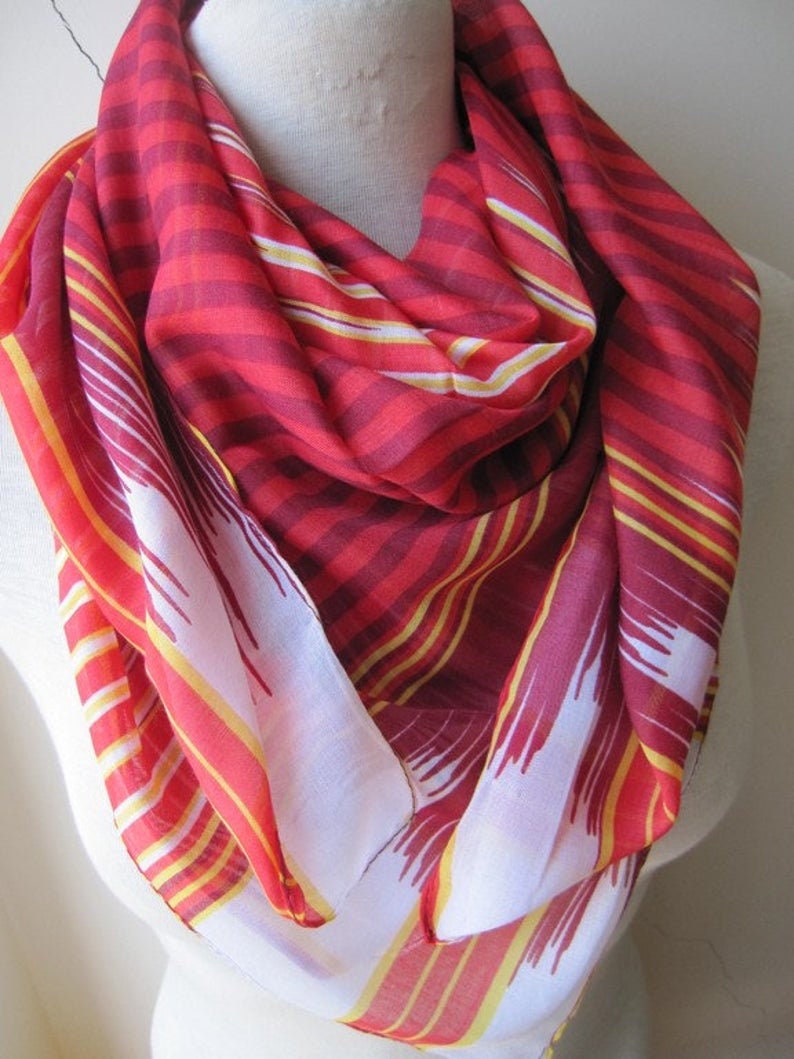 May include: A red and yellow striped scarf with a white and red geometric pattern. The scarf is folded and draped over a mannequin.