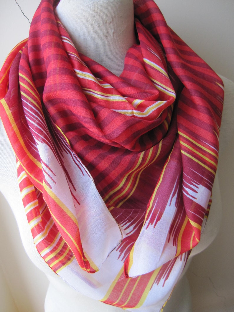 May include: A red and yellow striped scarf with a white background. The scarf is made of a lightweight fabric and has a fringed edge.